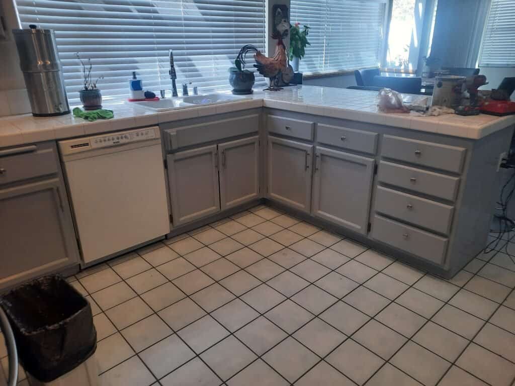 Gray kitchen cabinets being painted, featuring a tiled countertop, window blinds, and various kitchen appliances and decor elements.