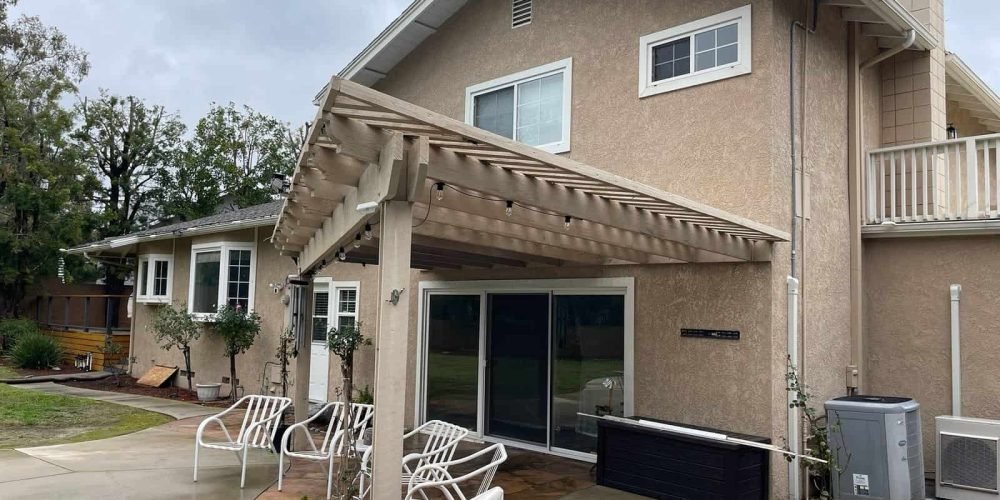 Bright two-story beige stucco house with a wooden pergola over a patio, outdoor seating, and lush landscaping, showcasing professional exterior painting and home improvement services.